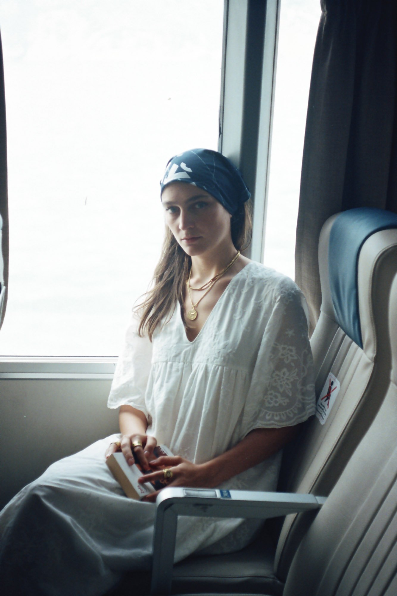 A woman wearing a white dress reading a book on a ferry boat seat