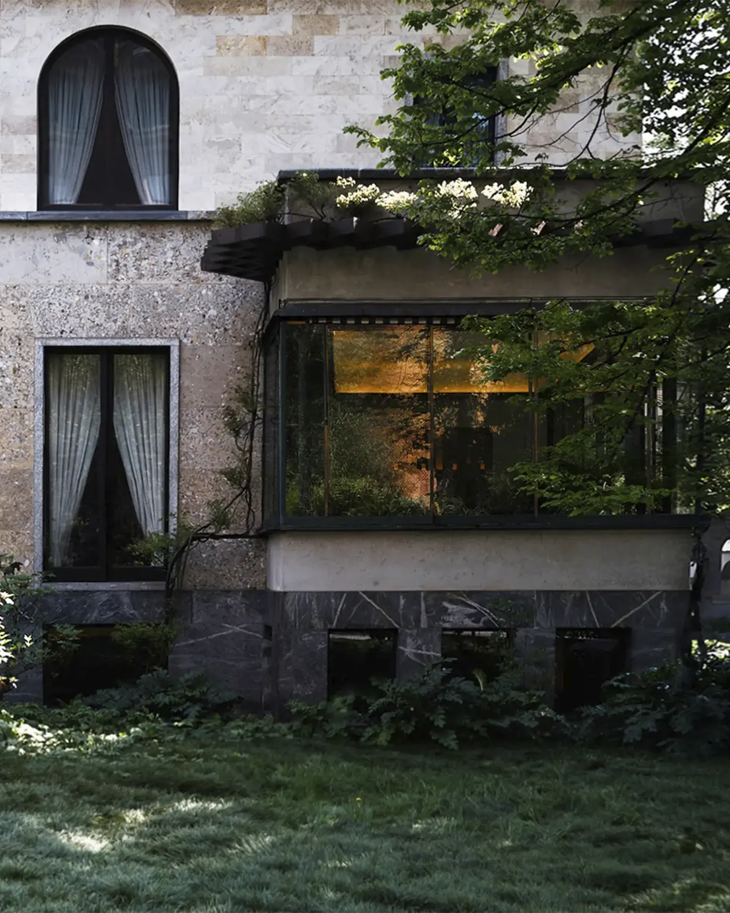 Exterior of Villa Necchi Campiglio with stone façade, arched windows, and glass garden room surrounded by greenery.