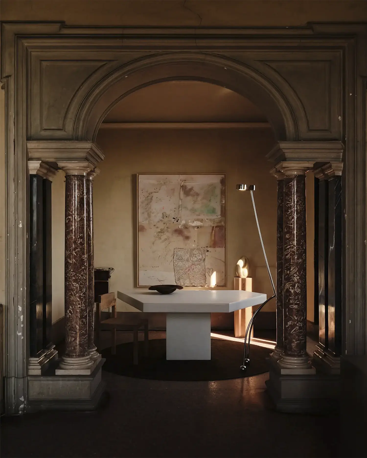 Installation view of a modern white table, sculptural objects, and wall art framed by a grand arch with marble columns in a historic villa.